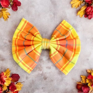 May include: A large, handmade hair bow with a plaid pattern in shades of yellow, orange, and a hint of blue. The bow is centered and has a classic bow shape. The background is a neutral grey with floral accents.