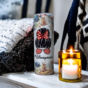 May include: A white tumbler with a colorful design featuring Disney characters and the text "#DisneyAdult". The tumbler has a black lid and a red bow with white polka dots.