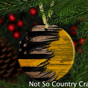 May include: A round, wooden Christmas ornament with a distressed American flag design. The flag is painted in black, white, and red with a thin yellow stripe across the middle. The ornament is hanging from a gold ribbon.