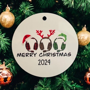 Könnte beinhalten: Runder weißer Weihnachtsbaumschmuck mit einem rot-schwarz-weiß karierten, einem leopardenfarbenen und einem grün-weiß gemusterten Headset mit Rentiergeweih und einem Weihnachtsmann-Hut. Der Baumschmuck trägt die Aufschrift "Frohe Weihnachten 2024".