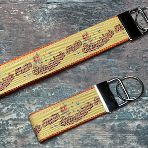 May include: Two yellow keychains with the text "Hello Sunshine" in a retro font. The keychains have a floral pattern and a silver metal clasp. The background is a dark, textured surface.