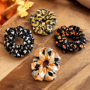 Op de afbeelding: Vier haar elastiekjes met verschillende Halloween-thema patronen. De elastiekjes zijn zwart met oranje en witte accenten. Een elastiekje heeft een patroon van snoep maïs, een ander heeft een patroon van geesten, een ander heeft een patroon van zonnebloemen en de laatste heeft een ruitpatroon.