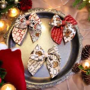 Op de afbeelding: Drie kerstthema haarstrikken met verschillende ontwerpen. De eerste strik is rood en wit met een Mickey Mouse-patroon en de tekst "Merry Christmas". De tweede strik is rood en wit met een peperkoekman-patroon en de tekst "Peperkoek en snoepstok". De derde strik is wit met een melk en koekjes-patroon en de tekst "Merry Christmas".