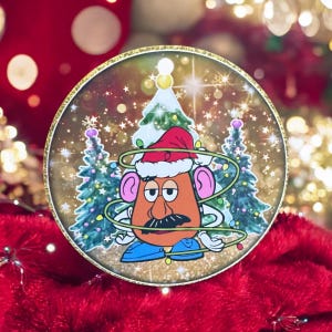 Puede incluir: Un adorno navideño redondo con Mr. Potato Head con un gorro de Papá Noel y enredado en luces navideñas. El adorno tiene un fondo de purpurina dorada y representa una escena de invierno nevada con dos árboles de Navidad.
