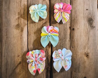 May include: Five colorful fabric hair bows with different patterns. The bows are arranged in a row on a wooden background. The bows are all made of soft fabric and have a variety of colors and patterns.