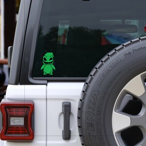 Pode incluir: Um Jeep branco com um adesivo de vinil verde de um personagem de desenho animado na janela traseira. O personagem tem um corpo verde, olhos pretos e uma boca preta. O adesivo est&aacute; na janela traseira do Jeep.