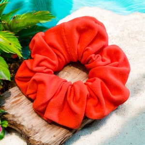 Puede incluir: Un scrunchie de color naranja brillante hecho de tela suave. El scrunchie tiene la forma de una flor grande y esponjosa.