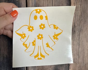May include: A white sticker featuring a yellow ghost design. The ghost is decorated with yellow flower accents. The sticker is on a white background, and the design is a whimsical, playful take on a Halloween theme.