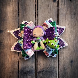 May include: Purple, green and white glitter bow with a purple and green character figurine of a girl with a green alien. The bow has a Toy Story theme.