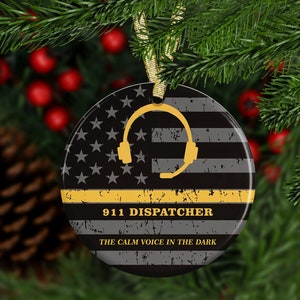 May include: A round, black and white ceramic ornament with a distressed American flag design. A yellow thin line and the words "911 Dispatcher" and "The Calm Voice in the Dark" are printed on the ornament.