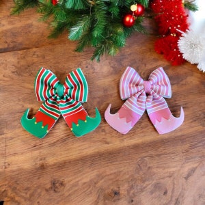 Puede incluir: Dos lazos para el pelo con temática navideña con diseños de zapatos de duende. Un lazo es rojo y verde con rayas blancas, el otro es rosa con rayas blancas.