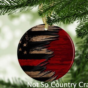 May include: A round, ceramic Christmas ornament with a distressed American flag design. The flag is red, white, and blue with a thin red line across the middle. The ornament is hanging from a gold string.