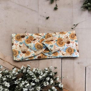 May include: A cream-colored headband with a repeating pattern of sunflowers and Highland cows. The headband is knotted at the top and has a soft, textured appearance. The design includes yellow, brown, and blue accents.
