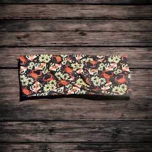 Może przedstawiać: Czarna opaska na głowę z powtarzającym się wzorem pomarańczowych krabów, żółtych stokrotek i małych czerwonych serc. Wzór zawiera również czarno-biały wzór w szachownicę. Opaska wykonana jest z miękkiego, elastycznego materiału.