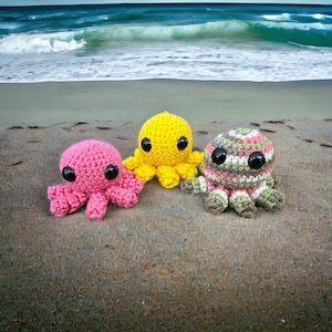 May include: Three crocheted octopus plushies in pink, yellow, and striped green and brown colors. The plushies have black button eyes and are sitting on a sandy beach.