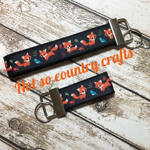 May include: Two black keychains with orange fox and blue bird designs. The keychains are on a wooden surface. The text "Not so country crafts" is visible in the image.