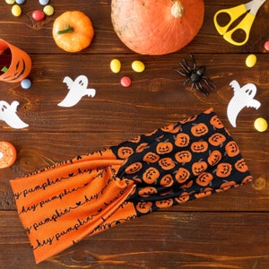 May include: A black and orange headband with a repeating pattern of pumpkins. The headband is made of fabric and has a knotted design. The headband is on a wooden surface with other Halloween decorations.