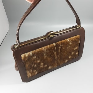 May include: A brown leather handbag with a flap closure and a gold-toned clasp. The front of the bag features a patch of brown and white fur.