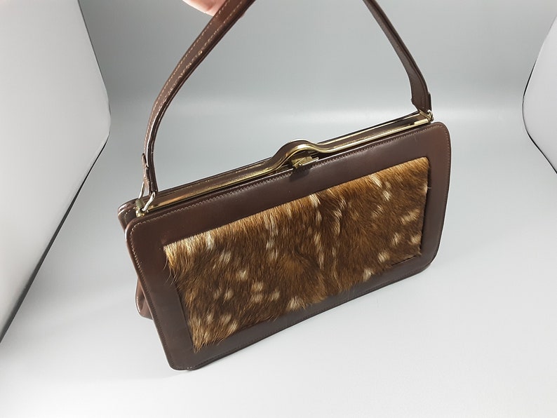 May include: A brown leather handbag with a flap closure and a gold-toned clasp. The front of the bag features a patch of brown and white fur.