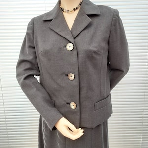 May include: A gray skirt suit with a blazer featuring a notched collar and three buttons. The blazer has a flap pocket on the left side. The skirt is knee-length.