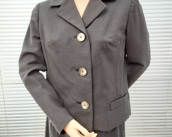 May include: A gray skirt suit with a blazer featuring a notched collar and three buttons. The blazer has a flap pocket on the left side. The skirt is knee-length.