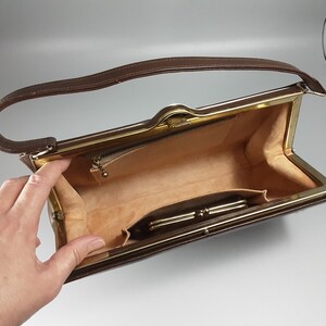 May include: A brown leather purse with a gold metal frame and a gold zipper. The purse is open and the inside is lined with a beige fabric. There is a small pocket on the inside of the purse.