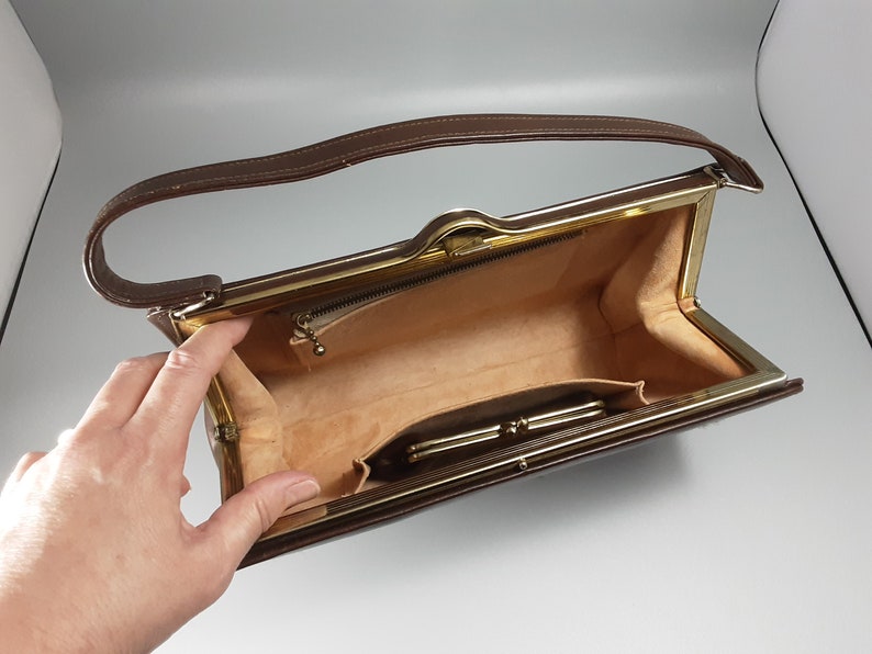 May include: A brown leather purse with a gold metal frame and a gold zipper. The purse is open and the inside is lined with a beige fabric. There is a small pocket on the inside of the purse.