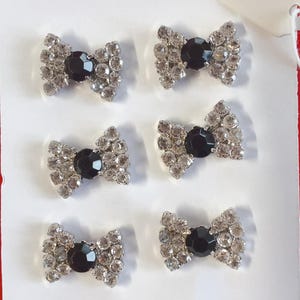 Puede incluir: Seis adornos decorativos en forma de lazo. Cada lazo presenta una gema negra central rodeada de brillantes pedrería transparente. Los lazos están dispuestos sobre una superficie blanca, con un borde rojo visible en el lateral.