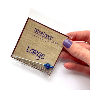May include: A close-up of a packaging for a large Ultra Punch The Precision Adjustable Needle. The packaging is brown with the text "Ultra Punch" and "Large" printed in blue. The packaging contains a silver needle with a blue tip and a silver spring.