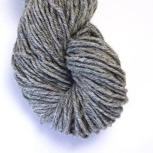 May include: A skein of gray yarn, made from wool, is shown in a close-up view. The yarn is tightly wound and has a soft, fuzzy texture.