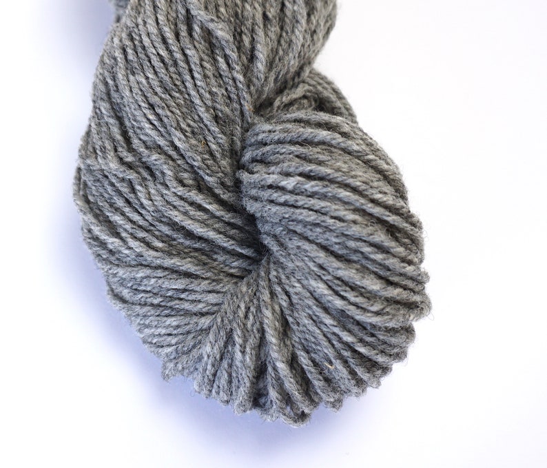 May include: A skein of gray yarn, made from wool, is shown in a close-up view. The yarn is tightly wound and has a soft, fuzzy texture.