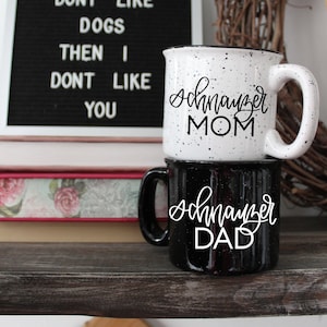 Op de afbeelding: Twee koffiemokken met zwarte letters op een wit en zwart gespikkelde achtergrond. De bovenste mok zegt "Schnauzer Mom" en de onderste mok zegt "Schnauzer Dad". De mokken staan op een houten plank met een boek en een krans erachter.