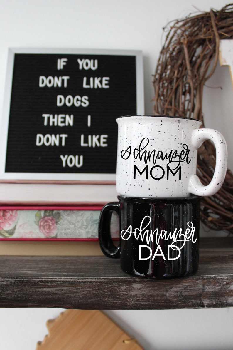 Op de afbeelding: Twee koffiemokken met zwarte letters op een wit en zwart gespikkelde achtergrond. De bovenste mok zegt "Schnauzer Mom" en de onderste mok zegt "Schnauzer Dad". De mokken staan op een houten plank met een boek en een krans erachter.