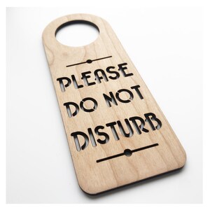 May include: A wooden door hanger with a hole at the top for hanging on a door knob. The hanger is light brown and has the text "PLEASE DO NOT DISTURB" in black lettering.