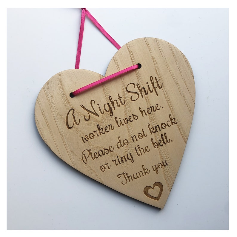 Puede incluir: Un letrero de madera en forma de coraz&oacute;n con el texto "A Night Shift worker lives here. Please do not knock or ring the bell. Thank you" grabado en &eacute;l. El letrero est&aacute; colgado de una cinta rosa.