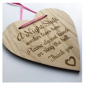 Puede incluir: Un letrero de madera en forma de coraz&oacute;n con el texto "A Night Shift worker lives here. Please do not knock or ring the bell. Thank you" grabado en &eacute;l. El letrero est&aacute; colgado de una cinta rosa.