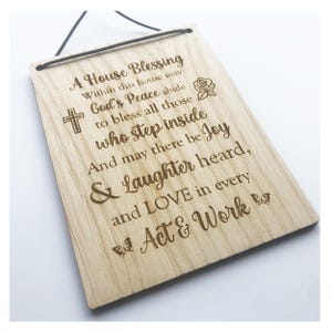 May include: A rectangular wooden plaque with a black hanging cord. The plaque has engraved text including "A House Blessing" and "God's Peace." Decorative elements include a cross, rose, and butterflies.