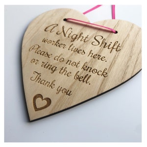 Puede incluir: Un letrero de madera en forma de coraz&oacute;n con el texto "A Night Shift worker lives here. Please do not knock or ring the bell. Thank you" grabado en &eacute;l. El letrero tiene una cinta rosa atada.