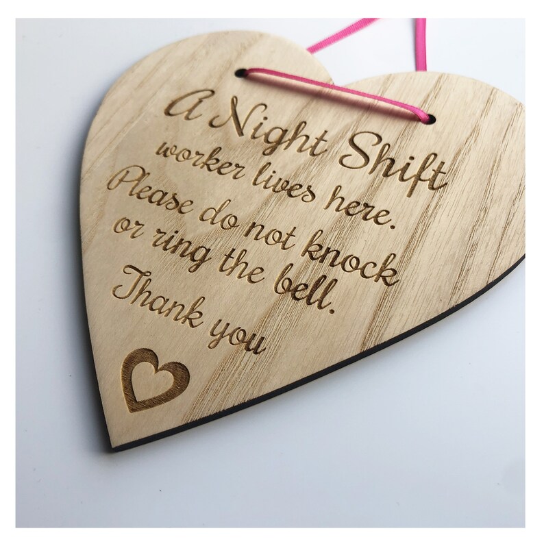 Puede incluir: Un letrero de madera en forma de coraz&oacute;n con el texto "A Night Shift worker lives here. Please do not knock or ring the bell. Thank you" grabado en &eacute;l. El letrero tiene una cinta rosa atada.