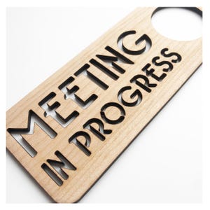 May include: A wooden door hanger with the text "Meeting in Progress" cut out in black. The hanger has a hole at the top for a door knob.