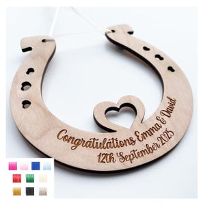 May include: Wooden horseshoe wedding decoration with the words "Congratulations Emma & David 12th September 2025" engraved. A heart shape is cut out of the wood. A white string is attached for hanging.