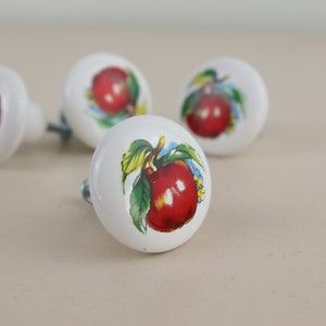 May include: Four white ceramic knobs with red apple designs. Each knob features a different apple design with green leaves and stems.