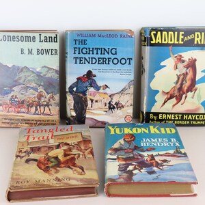 May include: A collection of vintage Western-themed books with colorful covers. Titles include "Lonesome Land," "The Fighting Tenderfoot," "Saddle and Ride," "Tangled Trail," and "The Yukon Kid." The books are arranged on a white surface.