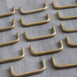 May include: A set of 14 gold-toned cabinet pulls, each with a simple, curved design. The pulls are arranged in rows on a light gray surface.