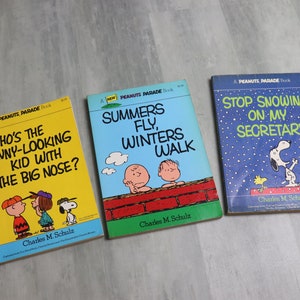 May include: Three vintage Peanuts paperback books with yellow, blue, and light blue covers. The titles are "Who's the Funny-Looking Kid with the Big Nose?", "Summers Fly, Winters Walk", and "Stop Snowing on My Secretary".