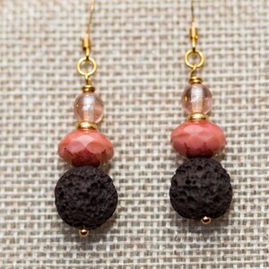 May include: A pair of earrings with a gold hook, three beads, and a lava rock. The beads are a light pink, a darker pink, and a clear glass bead. The lava rock is black and has a rough texture.