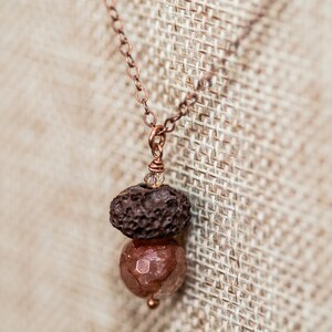 Puede incluir: Un collar de cadena de cobre con un colgante de piedra de lava marrón y una gema rosa.