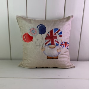 May include: Square cushion featuring a gnome illustration. The gnome is adorned with a Union Jack hat and flag, holding red and blue balloons and a medallion that reads "King Charles III Coronation". The cushion has a natural linen texture.