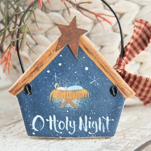 Può includere: Un ornamento natalizio a forma di casa, con sfondo blu, stelle bianche e la scritta "O Holy Night". Ha una cornice in legno marrone, una stella arrugginita e un nastro a righe rosse e bianche. L'ornamento è per la decorazione.