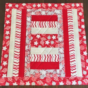 Peut inclure: Un chemin de table matelassé rouge et blanc avec un motif d'ornements de Noël. Le chemin de table a une bordure blanche et une bordure rayée rouge et blanche.